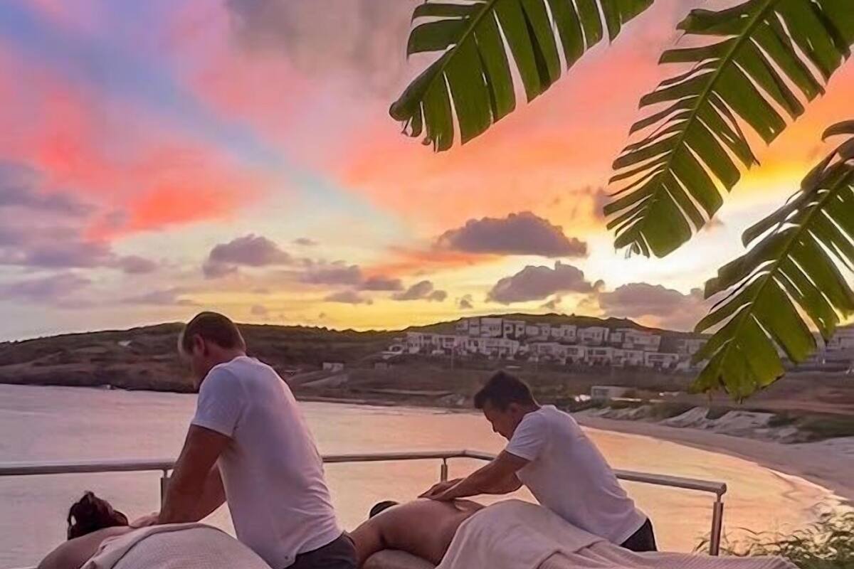 A massage therapy session with two clients on outdoor massage tables, overlooking a body of water and a distant shoreline during sunset.
