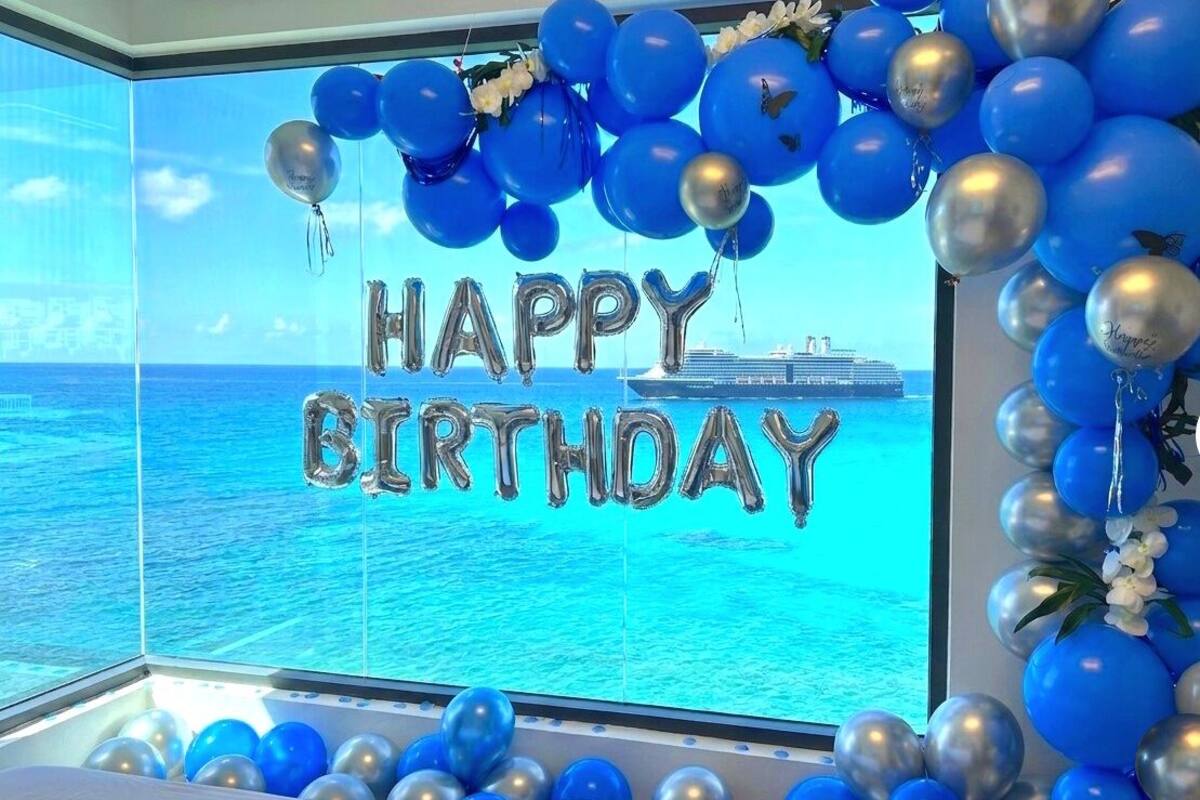 A room with a large window overlooking the ocean, decorated with blue and silver balloons and a 'Happy Birthday' sign.
