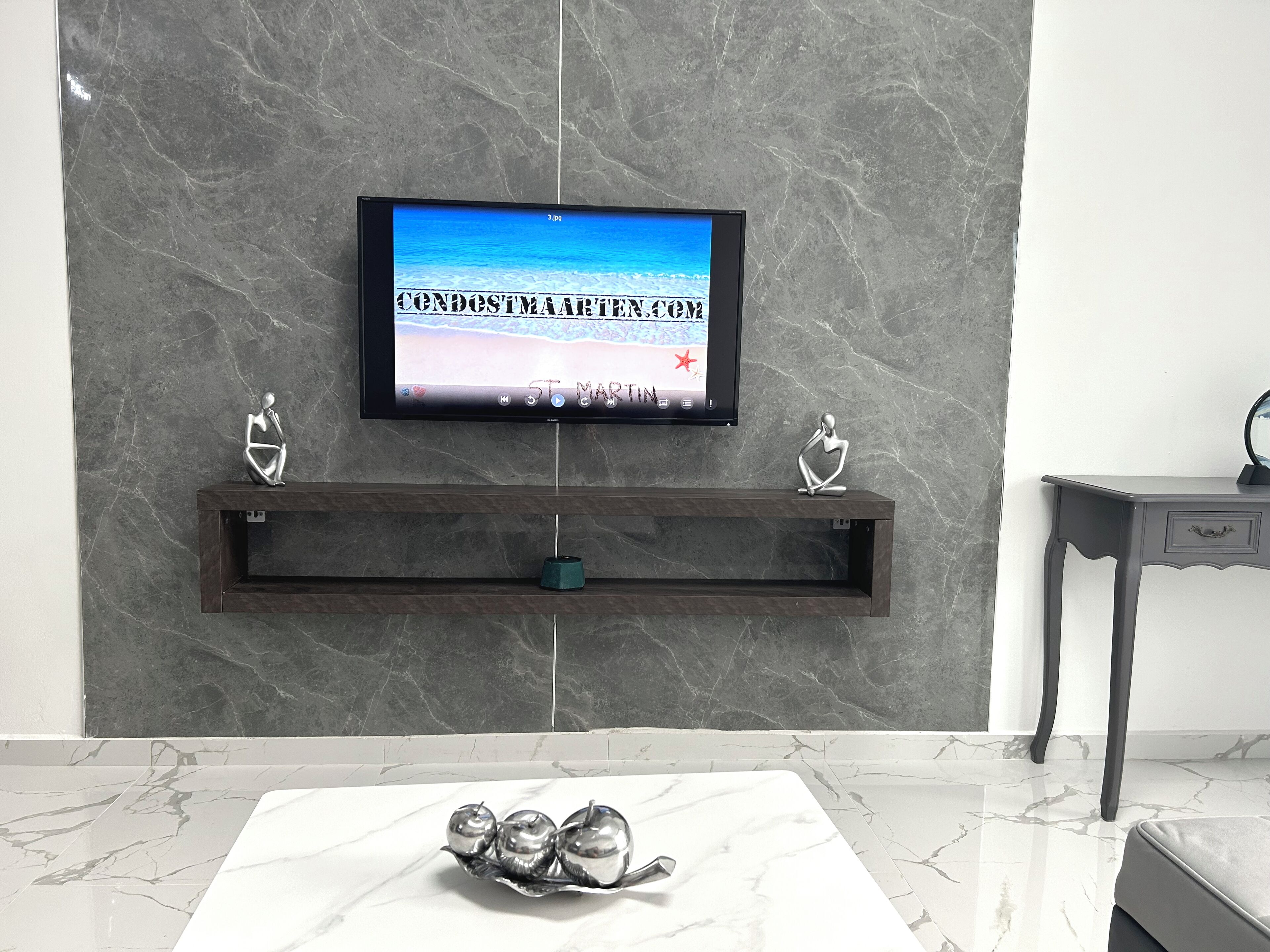 Una sala de estar moderna con un televisor de pantalla plana montado en una pared de mármol, un estante de madera oscura debajo y una mesa de centro de mármol con manzanas decorativas.