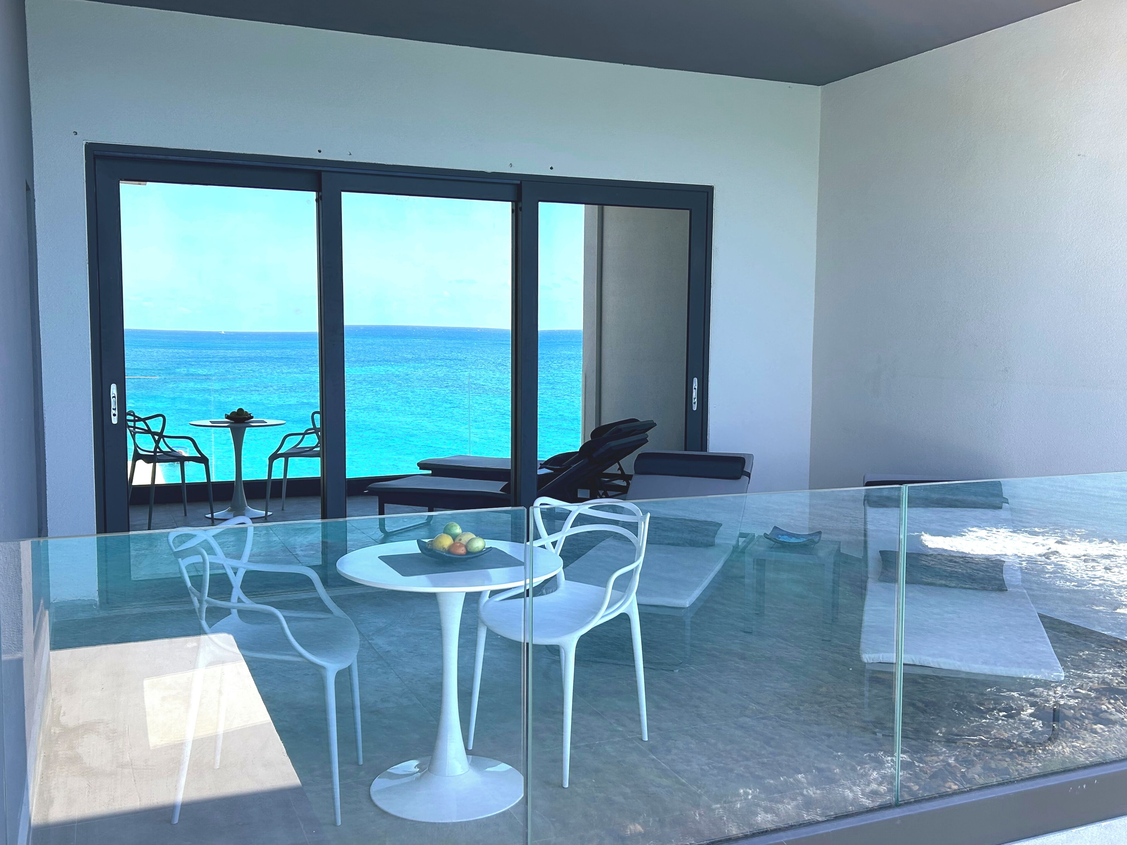 A modern balcony with a glass table and chairs, overlooking a clear blue sea.