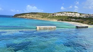 Een kustgezicht met helder turquoise water, een zandstrand en een heuvel met gebouwen.