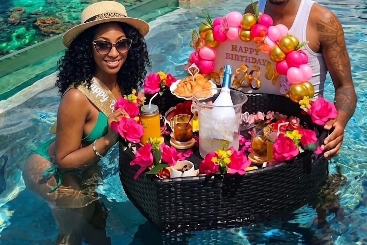A poolside area with clear water, a wicker basket with drinks and flowers, and a view of buildings and a hill in the background.