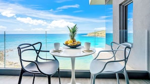 Een balkon met een tafel gedekt voor twee personen, met uitzicht op een strand en helderblauw water, met een verre kustlijn.
