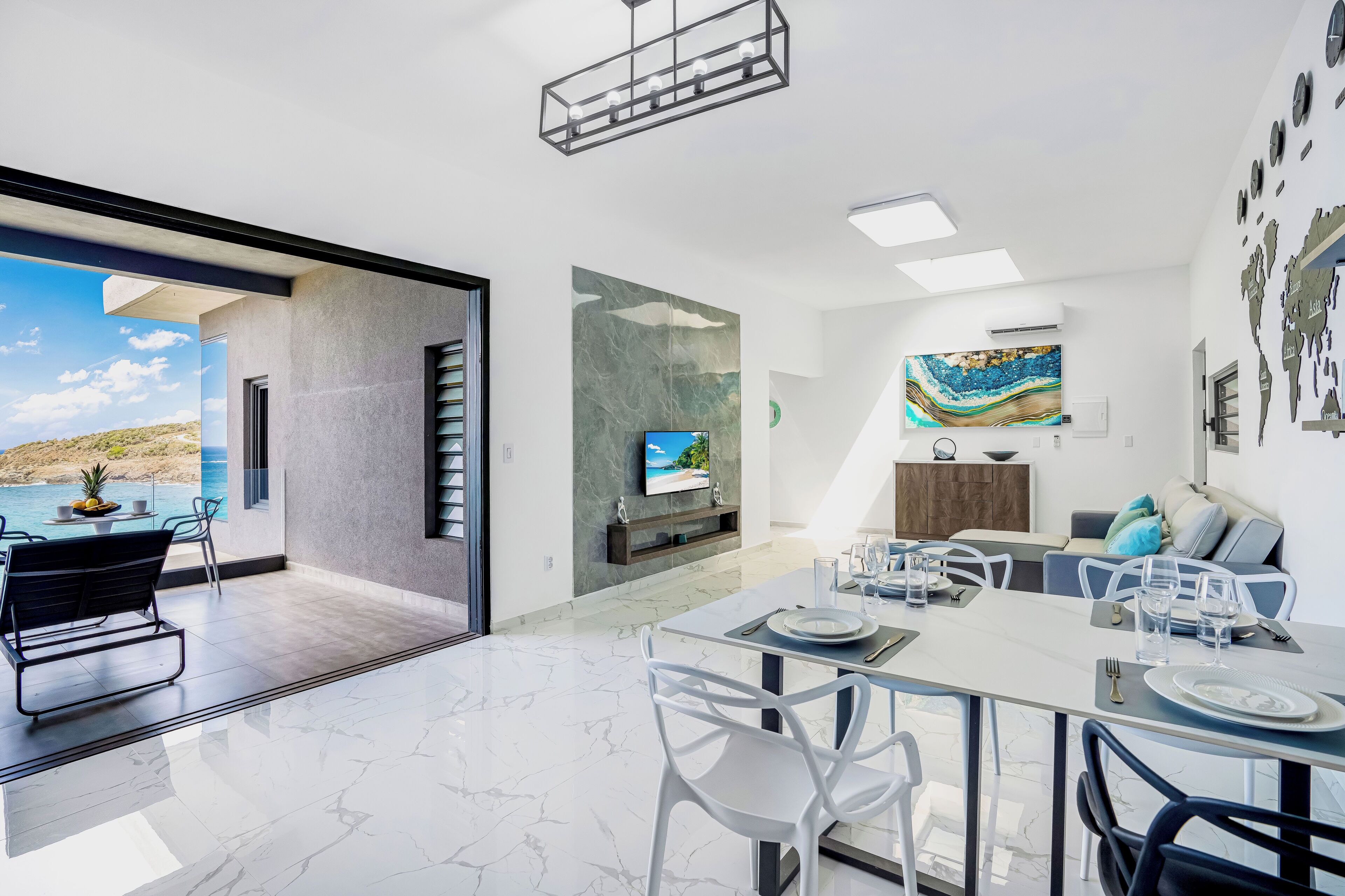 A modern dining area with a large table, white chairs, and a view of the sea.