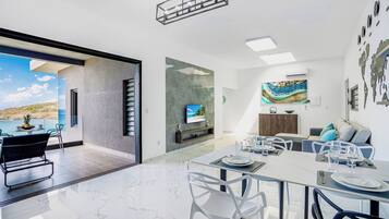 A modern dining area with a large table, white chairs, and a view of the sea.