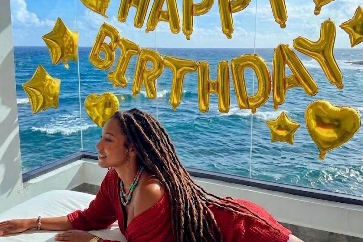 A room with a large window overlooking the ocean, decorated with gold balloons spelling 'HAPPY BIRTHDAY' and star-shaped balloons.