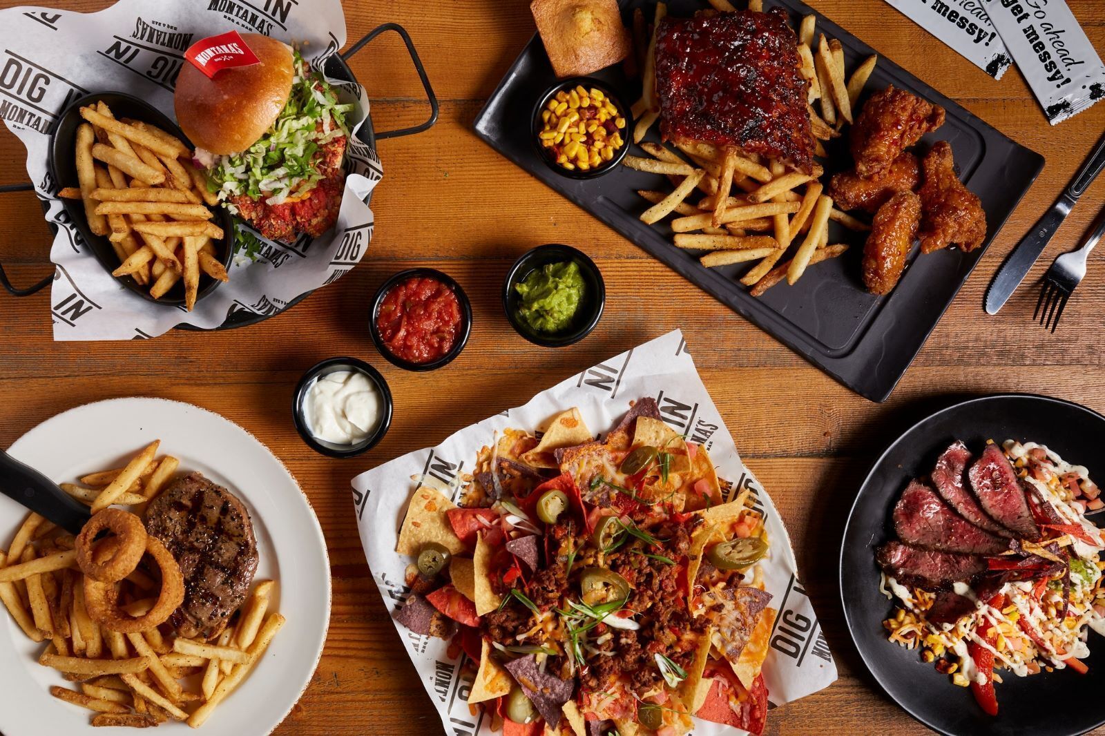 A variety of dishes including a burger, ribs, nachos, and steak, served on a wooden table with paper placemats.