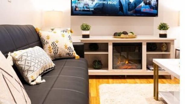 En moderne stue med et fladskærms-tv på væggen, en sort sofa med mønstrede puder, et træsofabord og en hvid, luvet måtte.