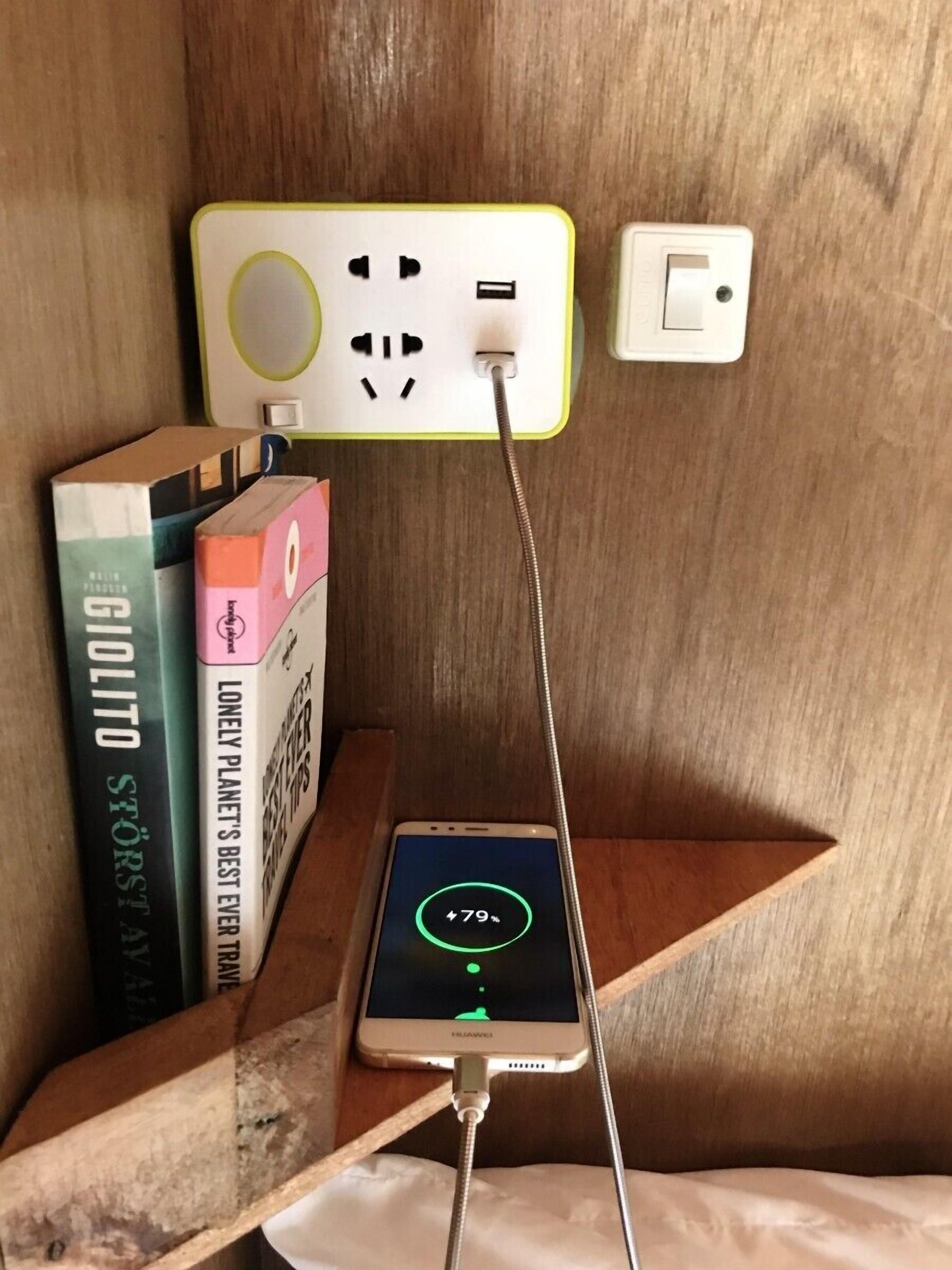 A smartphone charging on a wooden shelf with books, a power outlet, and a light switch.