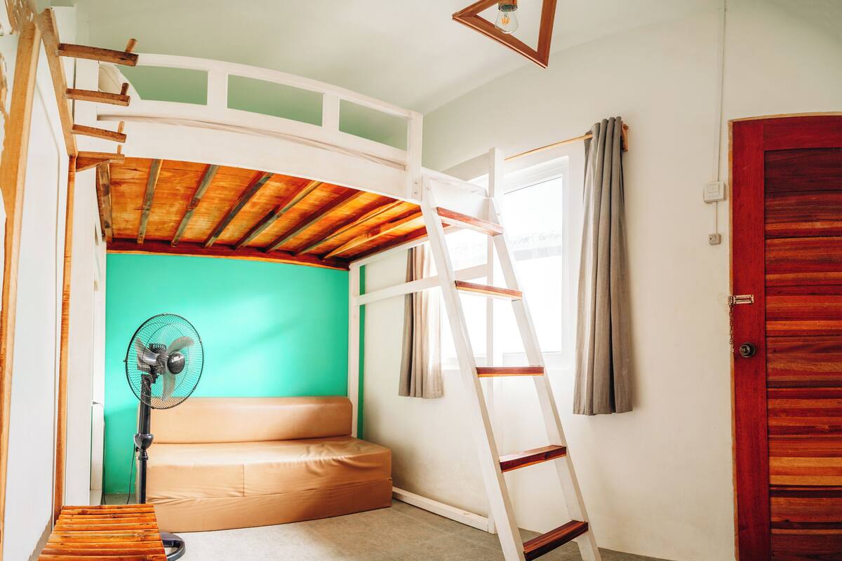 A room with a loft bed, a wooden bench, a ladder, a fan, and a window with curtains.
