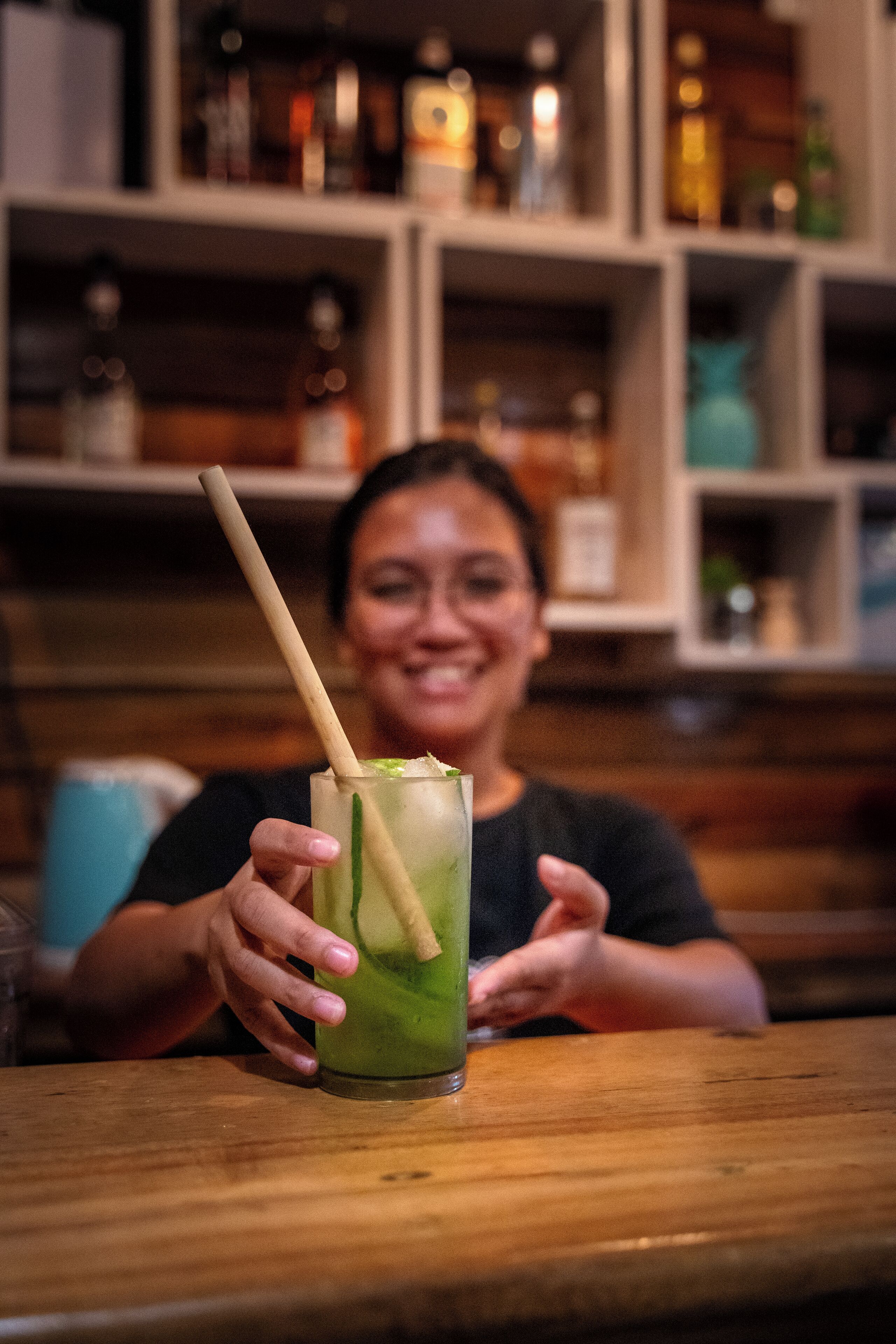 A person holding a green drink with a straw at a wooden table in a bar setting.