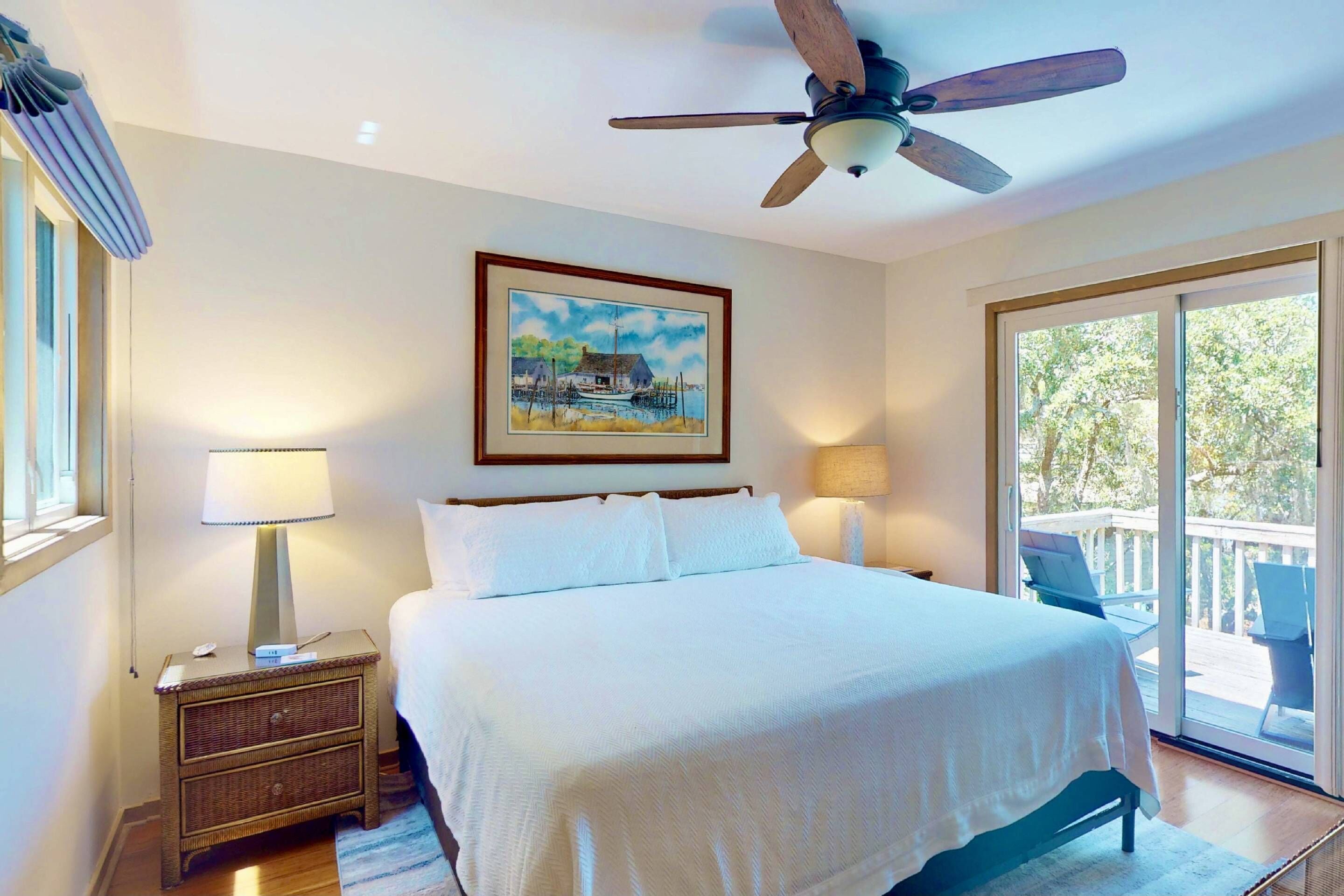 A bedroom with a bed, a ceiling fan, a nightstand with a lamp, a painting on the wall, and a sliding glass door leading to an outdoor area.