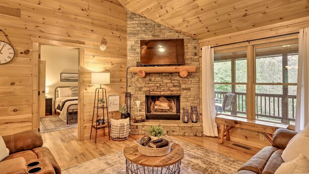 Interior acogedor de una cabaña de troncos con chimenea de piedra, mobiliario de madera y un ventanal grande con vistas al exterior.