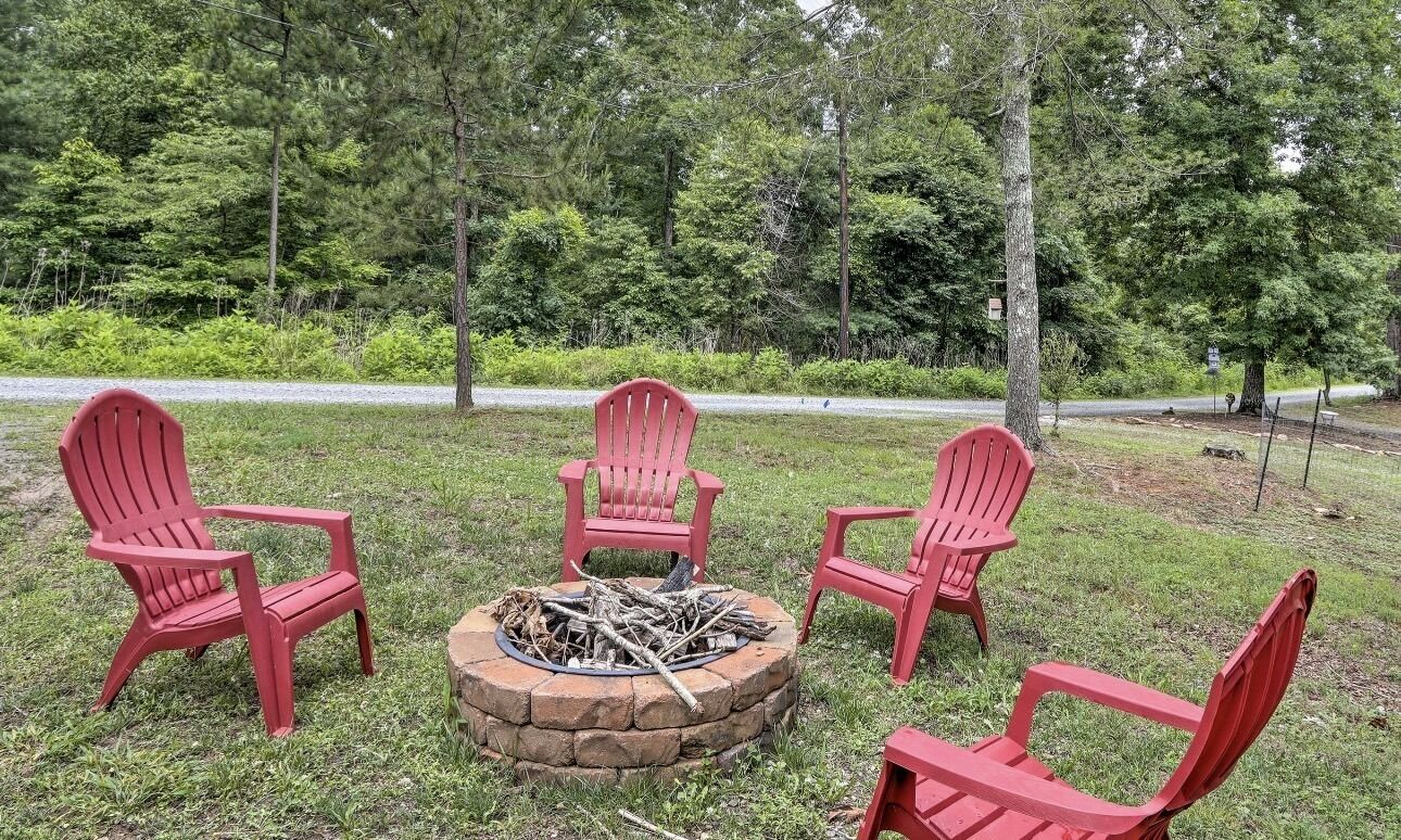 A fire pit with four red Adirondack chairs arranged around it in a grassy area with trees in the background.