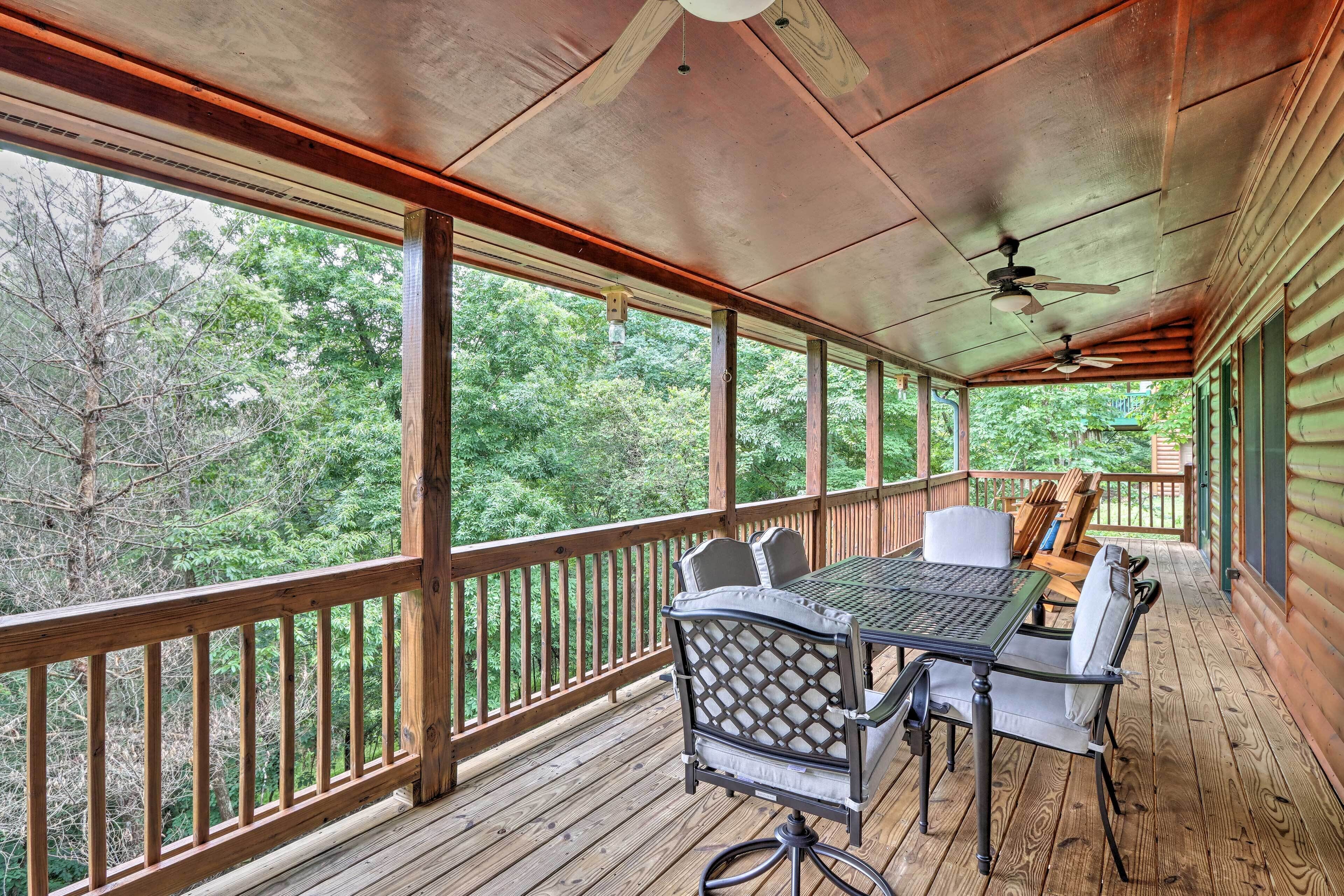 A covered outdoor deck with a table and chairs, surrounded by wooden railings and large windows offering a view of the forest.