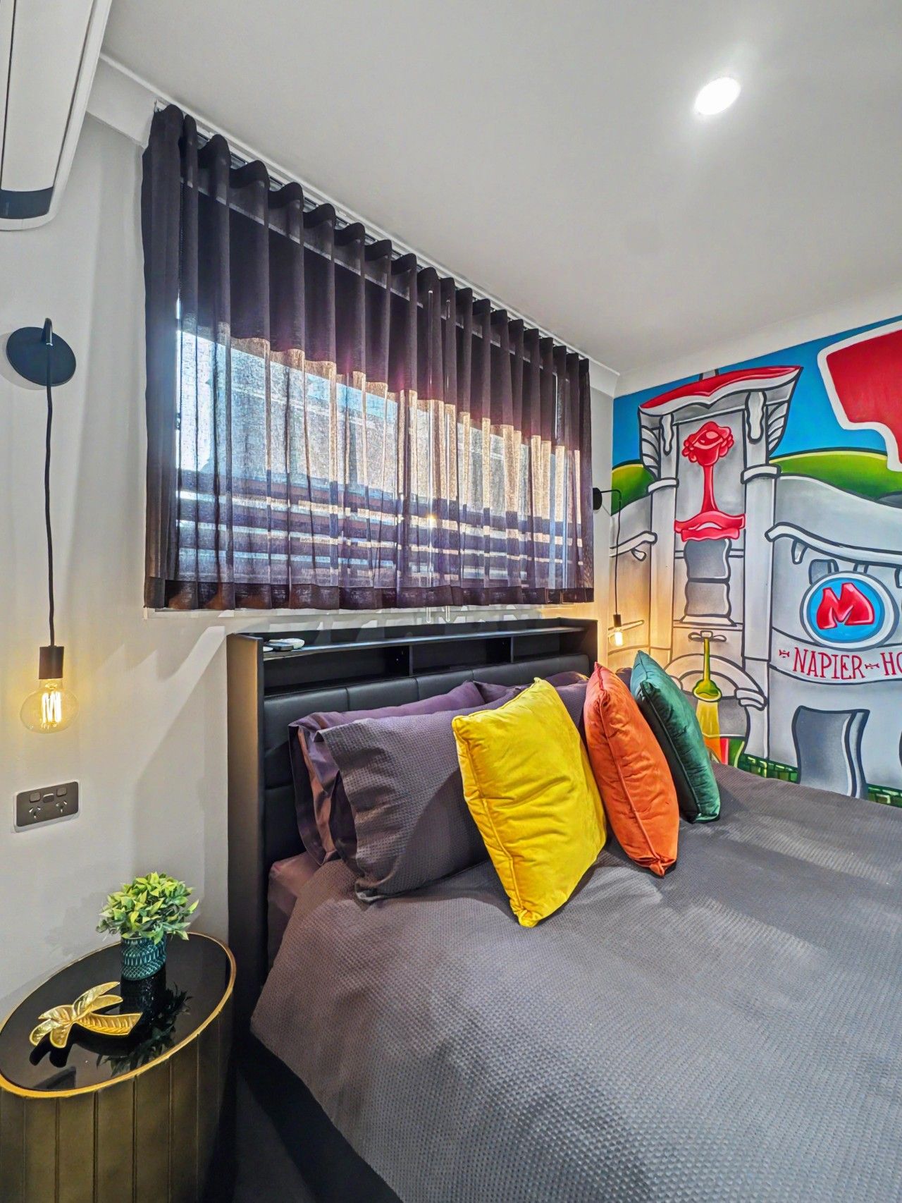 A modern bedroom with a bed, colorful pillows, a wall mural, and a window with curtains.