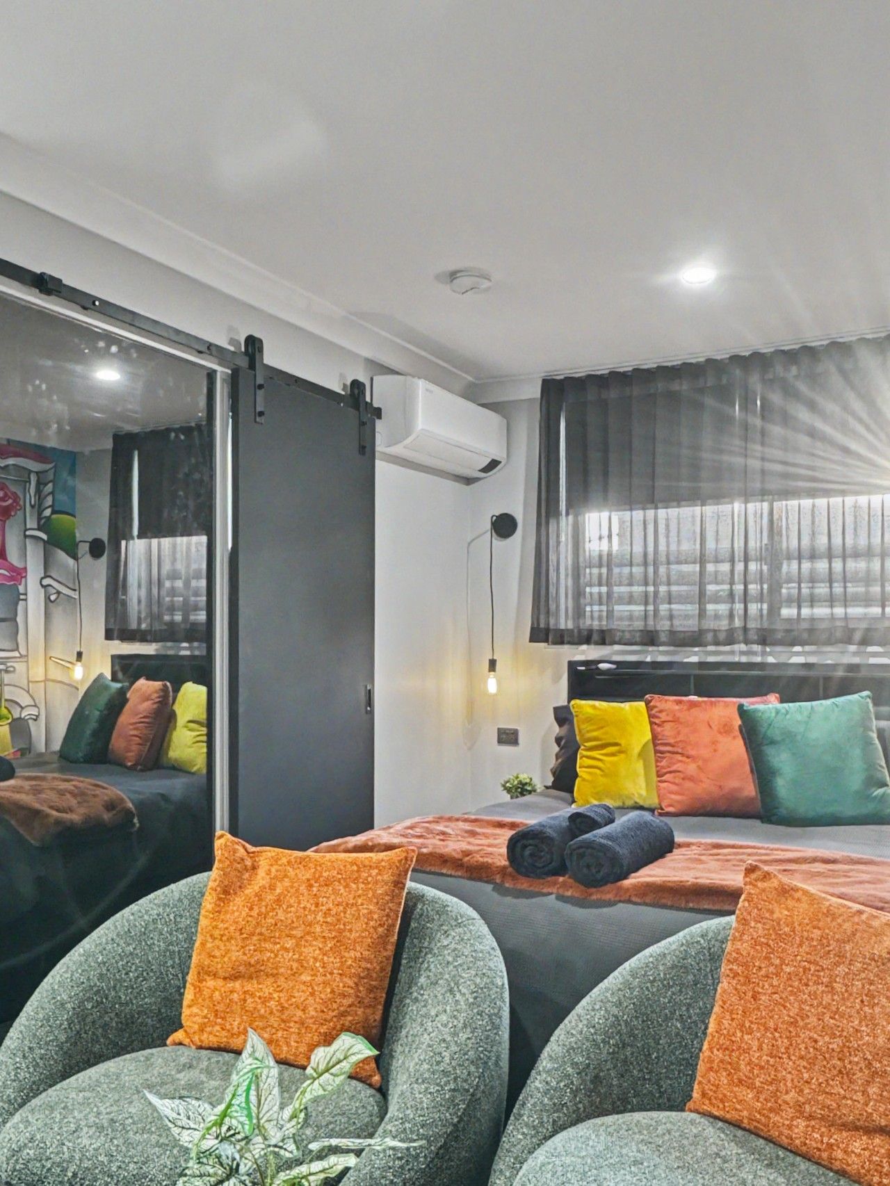 A modern bedroom with a bed, a seating area with chairs, a potted plant, and a large window with curtains.