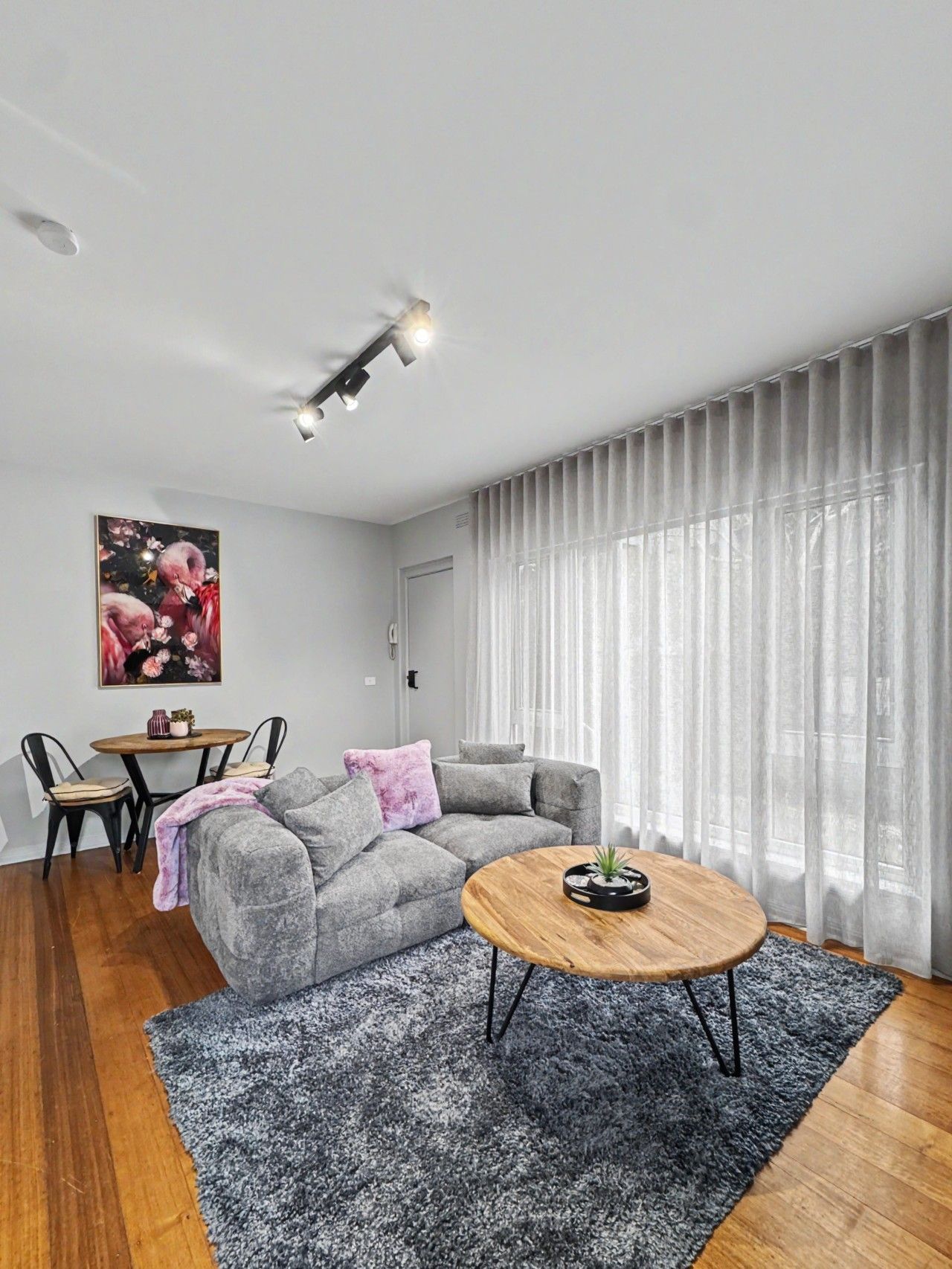 Una sala moderna con un sofá seccional gris, una mesa redonda, una mesa de comedor con sillas y un ventanal con cortinas traslúcidas.