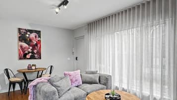Una sala moderna con un sofá seccional gris, una mesa redonda, una mesa de comedor con sillas y un ventanal con cortinas traslúcidas.