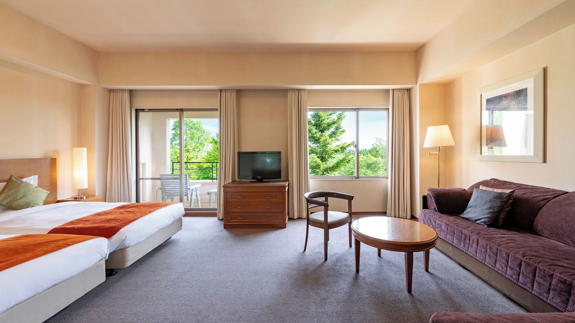 A hotel room with two beds, a TV, a sofa, a small table, and a balcony with a view of trees.