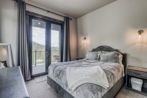 A bedroom with a large bed, a nightstand, a bedside table, and a view of the outdoors through a sliding door. - Whitefish Mountain Escape, 6 Bedroom Sleeps 14, Amazing Views! (Whitefish)