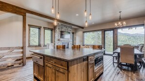 A spacious kitchen with a large island, wooden cabinets, and a stone fireplace. There are multiple windows offering views of the outdoors. - Whitefish Mountain Escape, 6 Bedroom Sleeps 14, Amazing Views! (Whitefish)