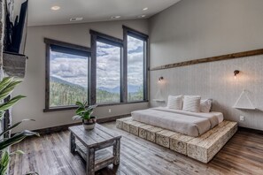 Une chambre avec un grand lit, un banc en bois et une vue sur les montagnes, visible à travers de grandes fenêtres.