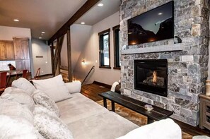 A living room with a stone fireplace, a large flat-screen TV mounted above it, and a cozy seating area with a sofa and cushions.