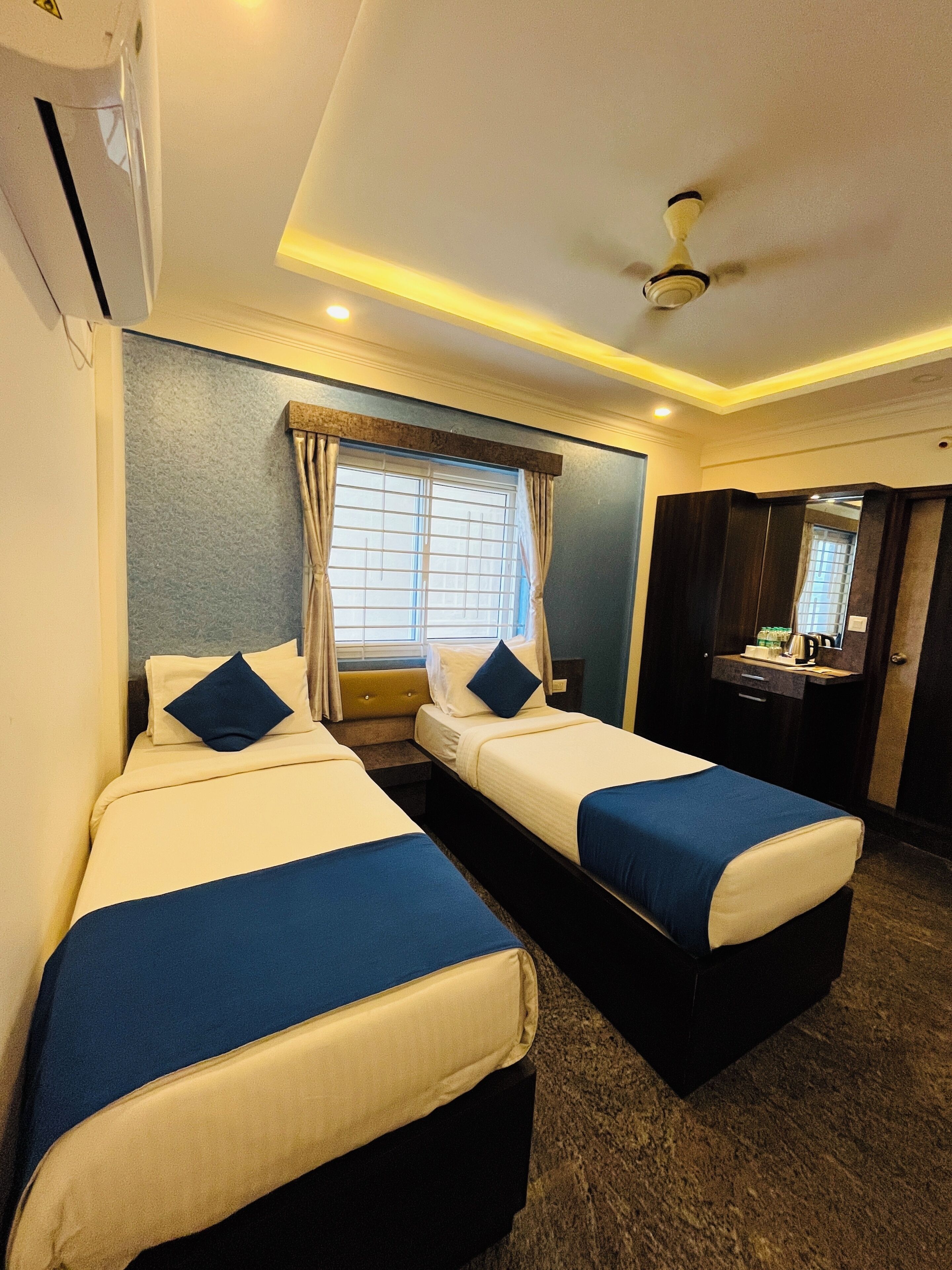 A hotel room with two beds, a ceiling fan, a window with curtains, a small desk with a mirror, and a bathroom with a sink and mirror.