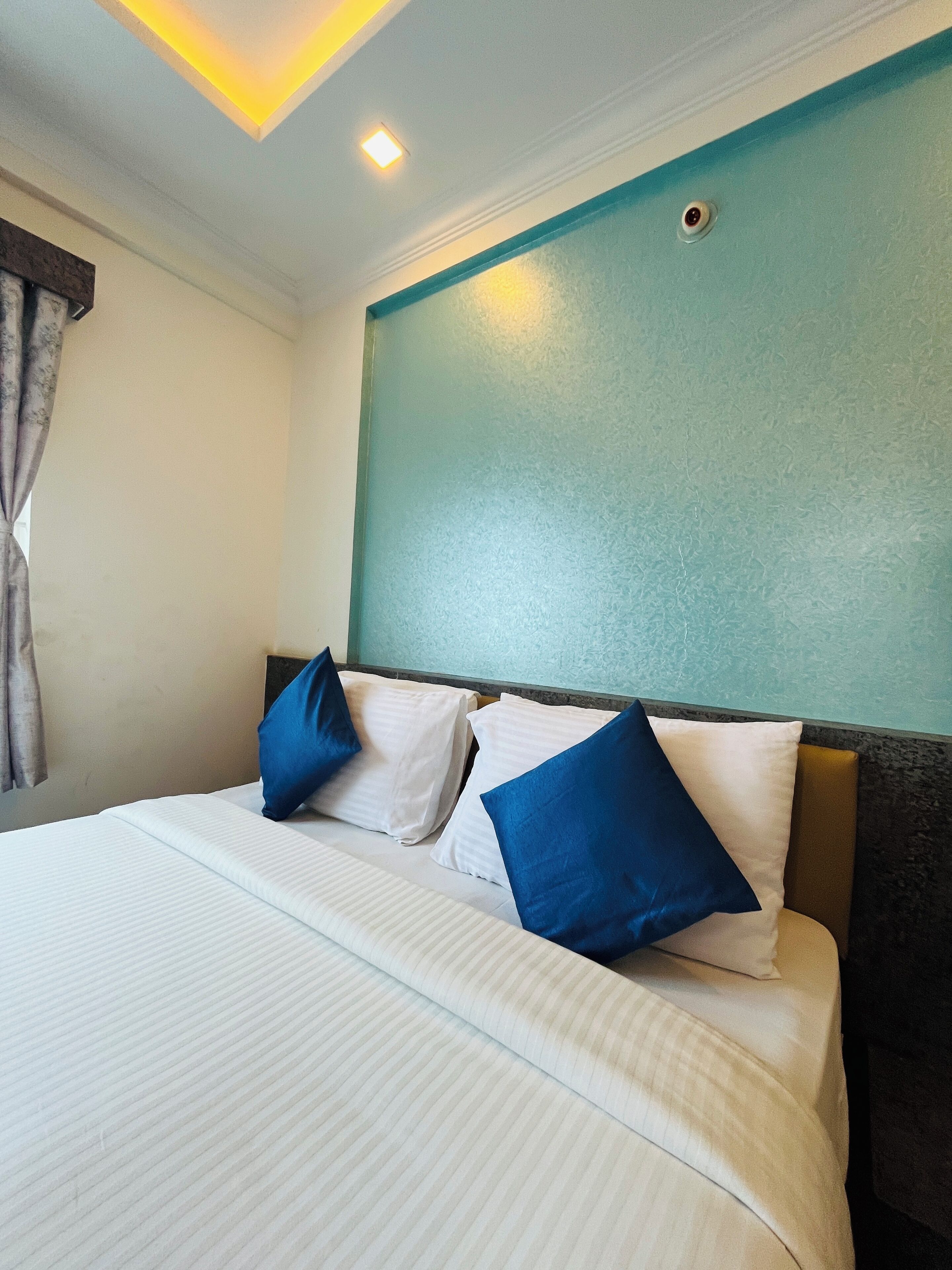 Een hotelkamer met een bed, kussens, een hoofdeinde, een raam met gordijnen en een plafondlamp.