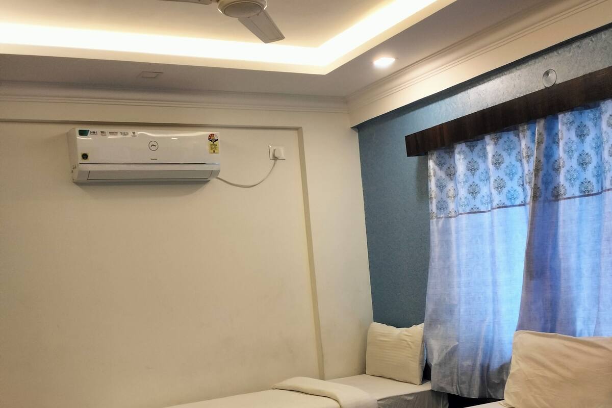 A room with two beds, a ceiling fan, an air conditioning unit, and a window with curtains.