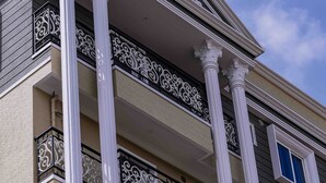 Un edificio de varios pisos con columnas blancas, balcones con barandillas negras ornamentadas y un cielo azul despejado al fondo.
