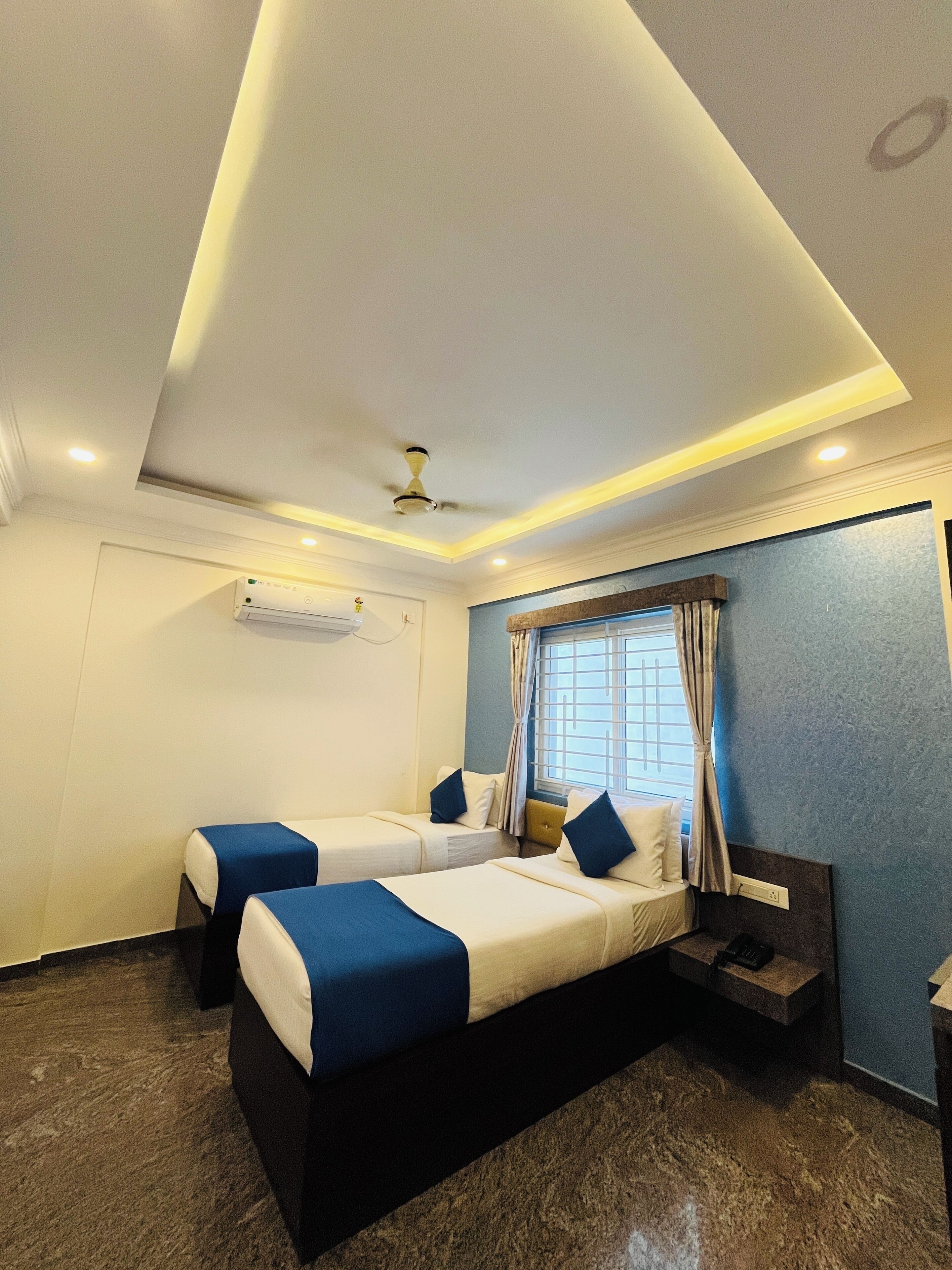 A hotel room with two beds, a ceiling fan, an air conditioning unit, a window with curtains, and a small table with a phone.