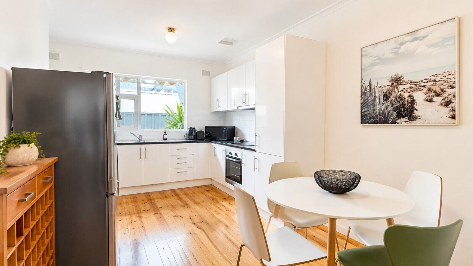 A modern kitchen with white cabinets, a black refrigerator, a dining table with chairs, and a painting on the wall.