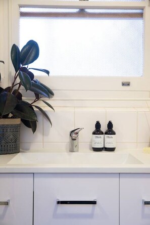 Une salle de bain avec un lavabo blanc, deux bouteilles d’huile d’olive, une plante en pot et une fenêtre avec des stores.