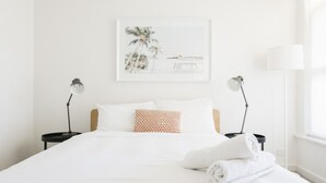 A neatly made bed with white linens and a folded towel, two bedside lamps, a framed picture on the wall, and a window on the right side. - 2 BR Apt | Free Parking & Wi-Fi Near Glenelg Beach (Glenelg East)