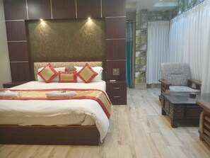 A bedroom with a large bed, a nightstand, a chair, and a small table. - The Brahmaputra Hotel (Dhubri)