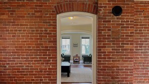 Une pièce aux murs de briques et au plafond en bois, une porte-fenêtre donnant sur une autre pièce où se trouvent une table et des chaises, et un petit objet noir accroché au mur.