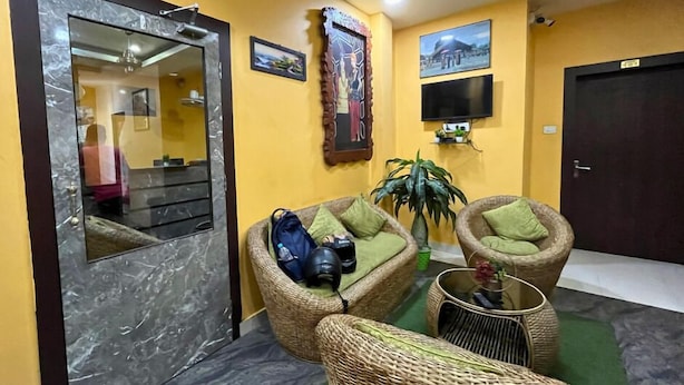Un área de recepción con sillones de mimbre, un espejo grande, una planta en maceta y un televisor montado en la pared.