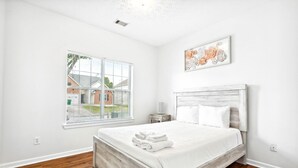 Un dormitorio con cama de madera, paredes blancas, una ventana con persianas y una obra de arte enmarcada en la pared.