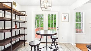 Una habitación con una mesa redonda y dos taburetes, una estantería, una puerta con vistas y una lámpara colgante.