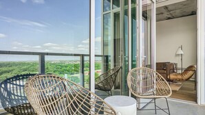A balcony with wicker chairs and a table, a glass door leading to a room with a sofa and a lamp, and a view of a green landscape. - Modern Downtown Condo*Rooftop pool*Top Amenities (Austin)
