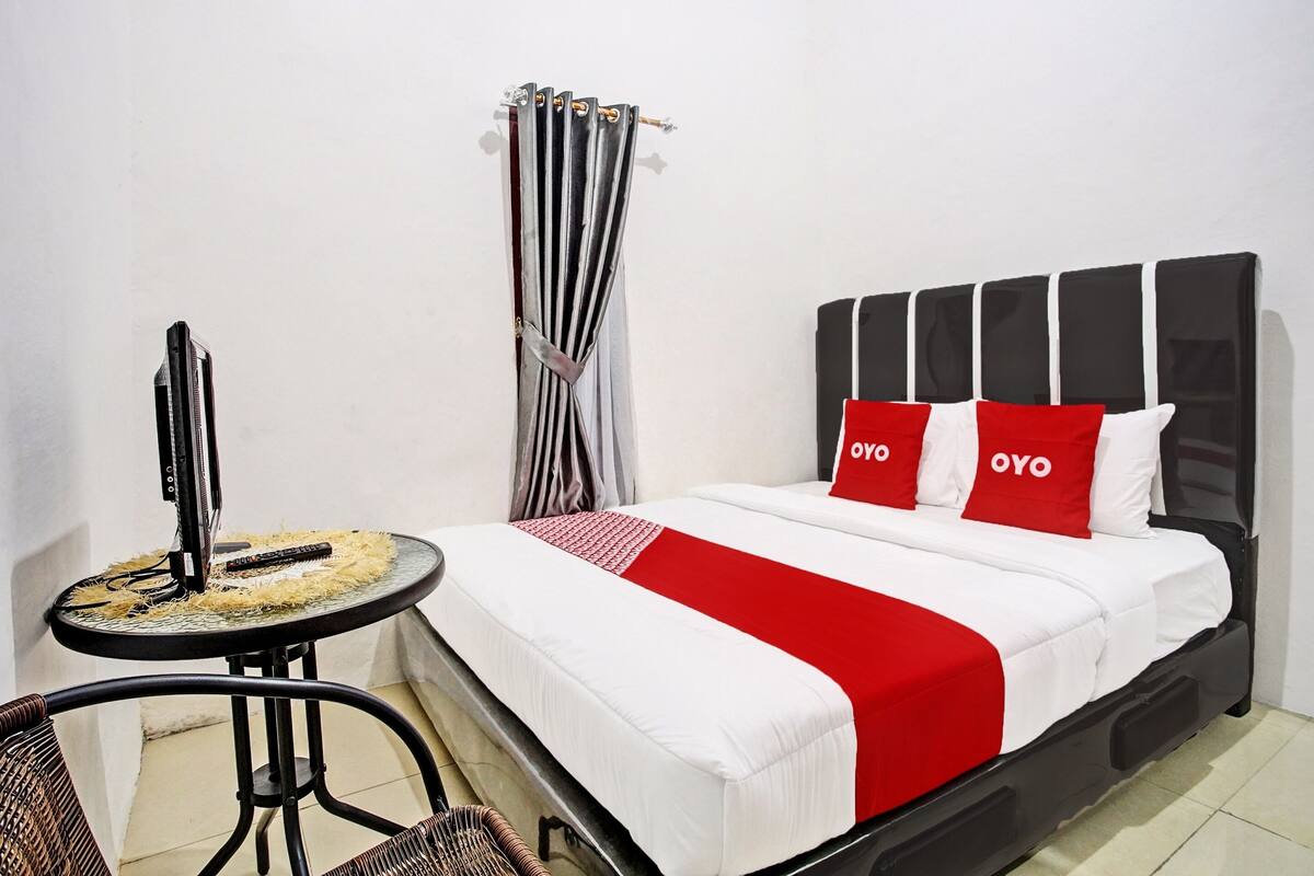 A bed with a black headboard, white bedding, and red accents. There is a small round table with a chair beside the bed, and a curtain is visible in the background.