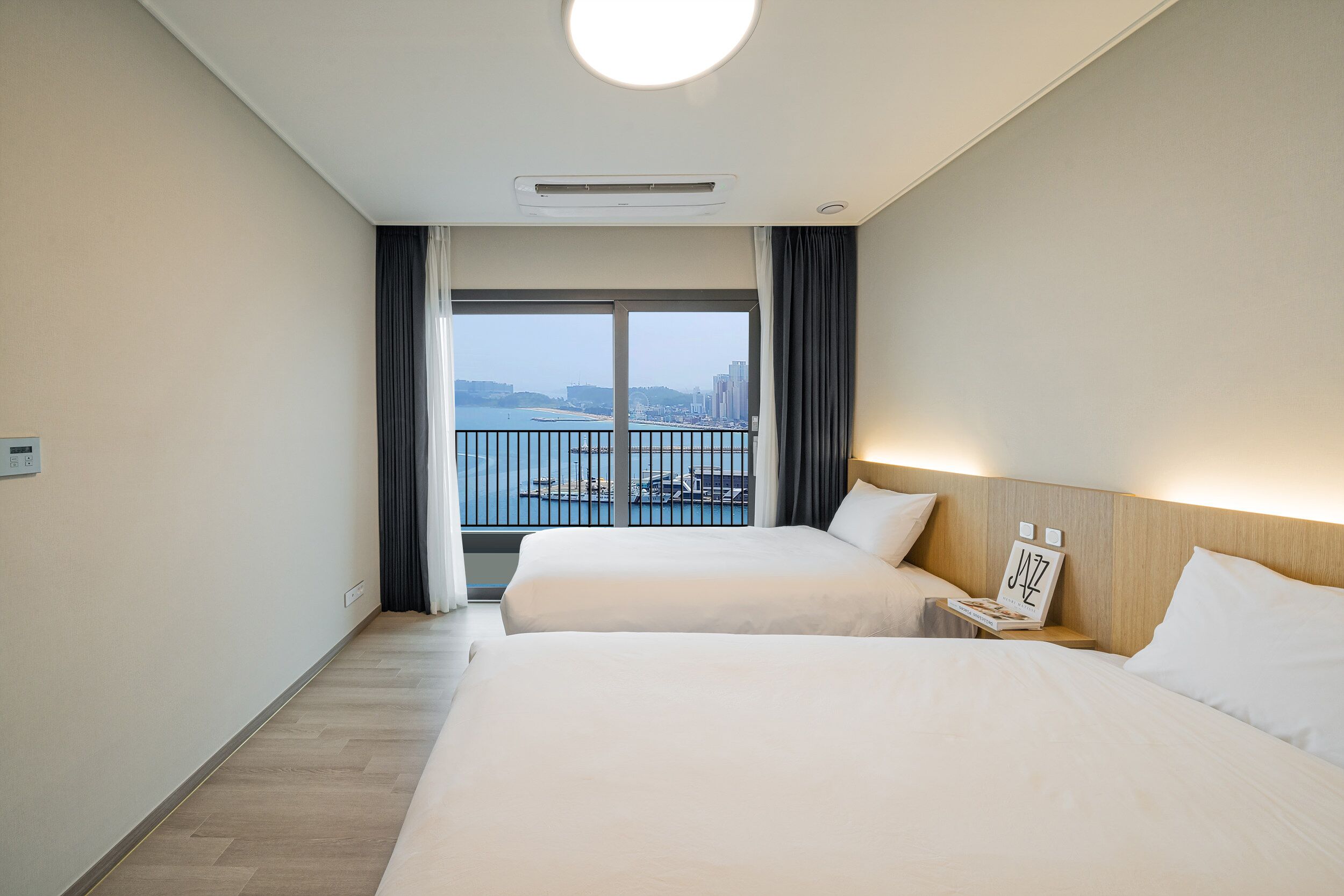 A hotel room with two beds, a view of the city through large windows, and a balcony with a railing.
