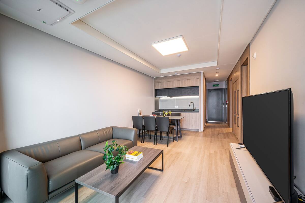 Una sala de estar moderna con un sofá, una mesa baja con un libro y una planta, un televisor de pantalla plana y una cocina al fondo.
