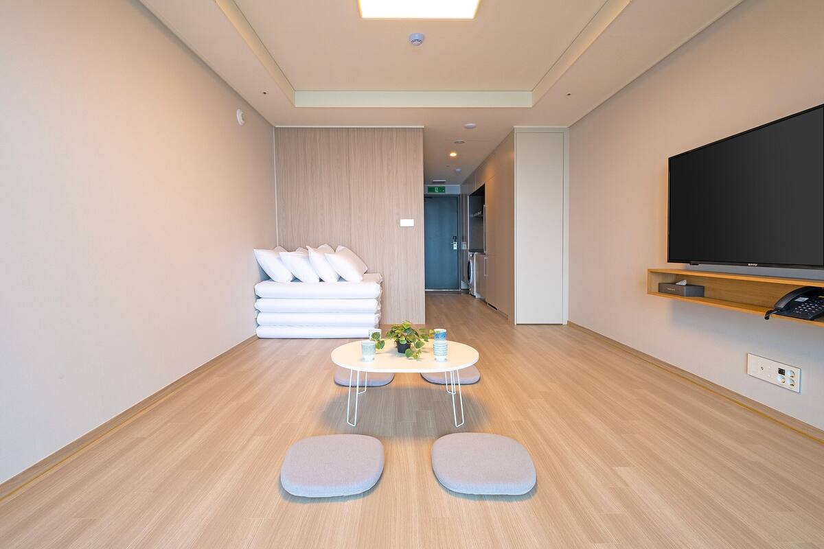 Una habitación de hotel moderna con suelo de madera, un televisor de pantalla plana en la pared, una pequeña mesa redonda con una planta y una zona de estar con cojines.