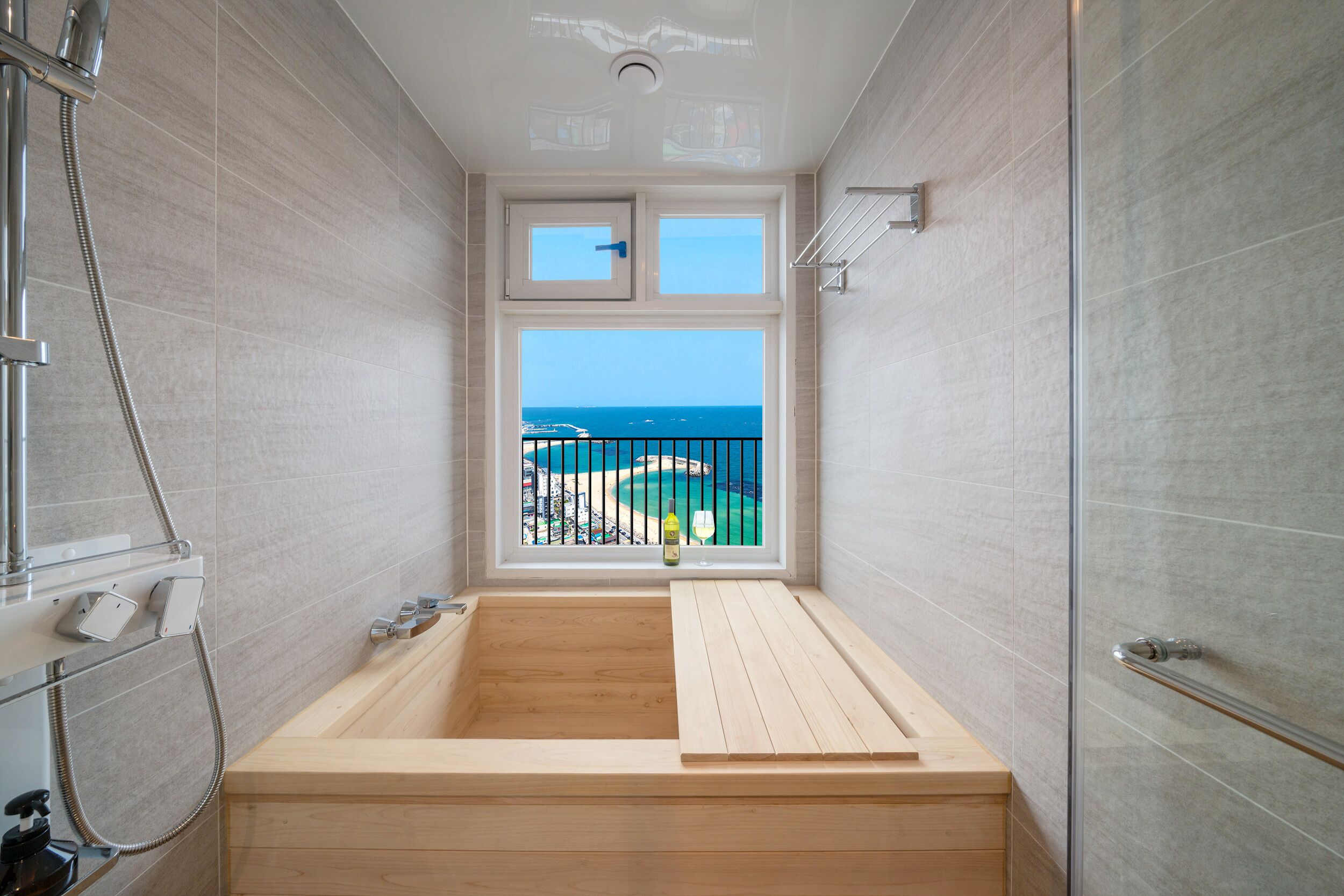 A modern bathroom with a wooden bathtub, a shower, and a window offering a view of the beach.