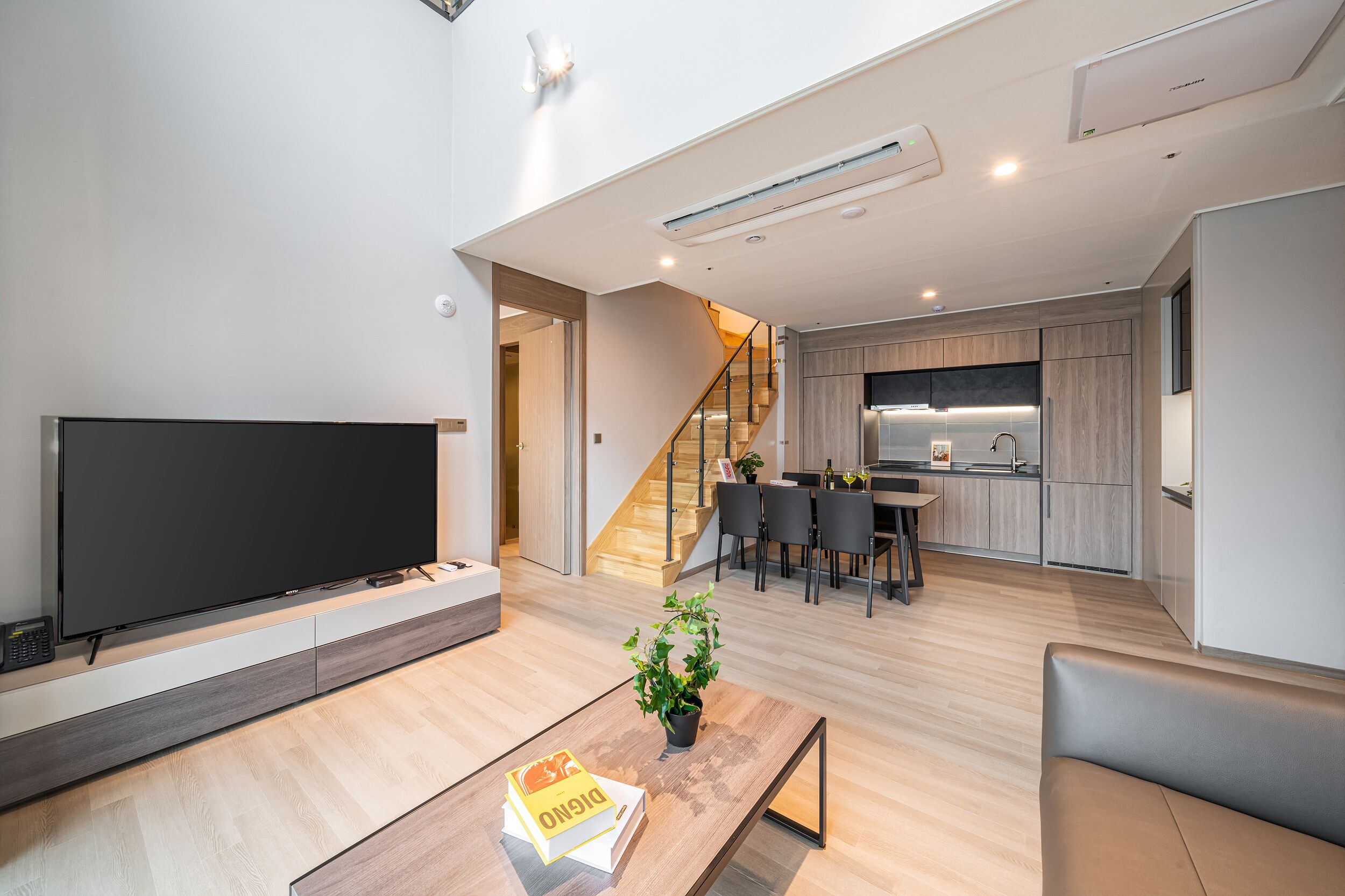 Een moderne woonkamer met een flatscreen televisie, een bijzettafeltje met een boek en een plant, en een trap naar een keuken.