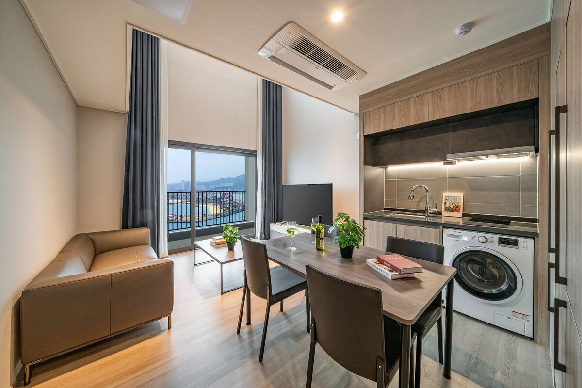 Una cocina moderna con lavadora y secadora integradas, una zona de comedor con mesa y sillas, y un amplio ventanal con vistas a la ciudad.