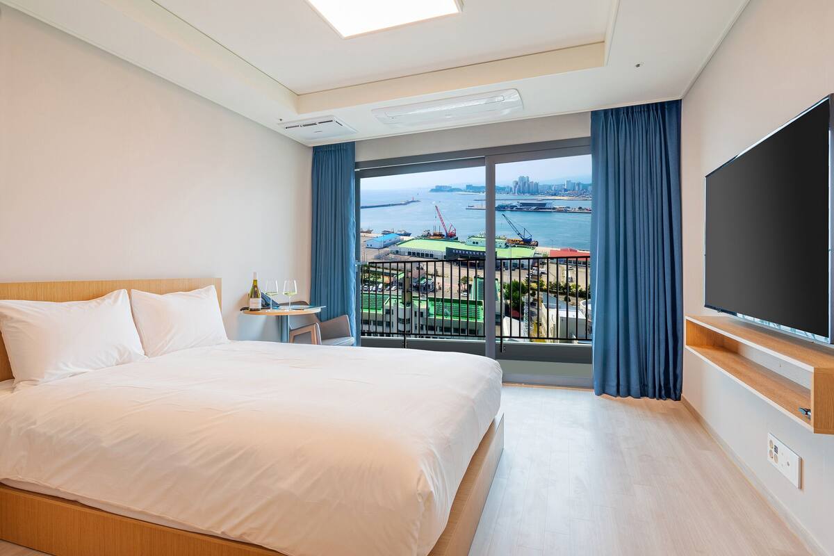 Habitación de hotel con una cama grande, televisión y balcón con vista al agua y al horizonte de la ciudad.